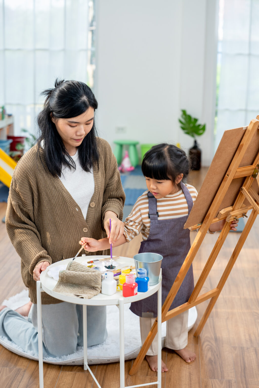 Young asian mom guides preschool daughter selecting paint and brush from supply table near easel. Single mother supports creative play, color learning, fine motor skill, bonding, patience, care home