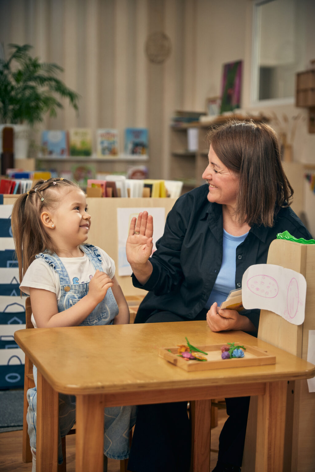 Smiling educator giving high five to happy girl after preschool learning activity. Concept of positive reinforcement, childcare confidence, emotional support, progress, and joyful education.