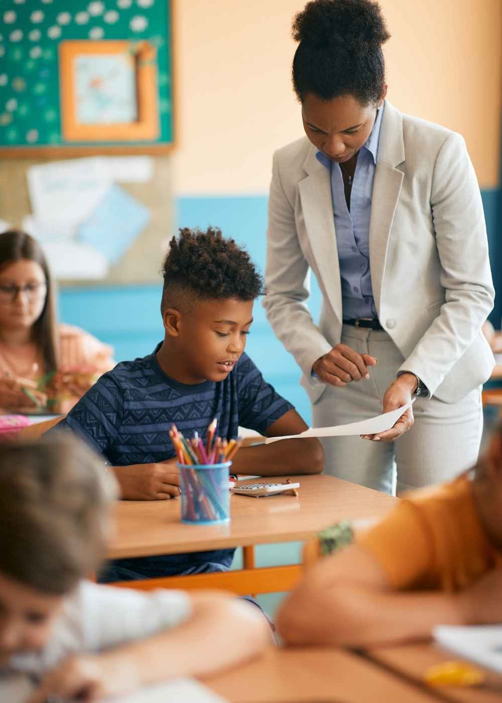 Teacher helping a young boy with a classroom assignment while other children work at their desks.