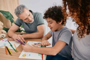 Child drawing with colored pencils while two adults provide encouragement and support.